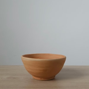 Puede incluir: Un cuenco de terracota de color naranja-marrón, de forma redondeada y con una amplia abertura. Descansa sobre una superficie de madera de color claro. El fondo es de un gris claro liso.