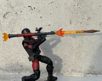 May include: A black and red action figure holding a black and orange rocket launcher with a bright orange flame effect. The figure is kneeling on a black base.