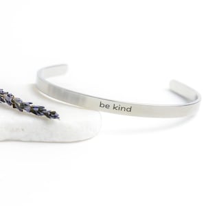 May include: A silver bangle bracelet with the words "be kind" engraved on it. The bracelet is resting on a white stone with a dried lavender sprig.