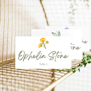Puede incluir: Tarjetas de mesa blancas con diseños florales y caligrafía elegante. Las tarjetas presentan ilustraciones de acuarela de flores amarillas, hojas verdes y ramitas de lavanda. La tarjeta frontal dice "Ophelia Stone" y "Table 2".
