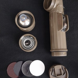 Puede incluir: Una linterna desmontada con un cuerpo verde y un reflector plateado. La linterna está compuesta por cinco partes: el cuerpo, el reflector, la lente, el porta pilas y el interruptor. La linterna está desmontada, con las piezas dispuestas sobre una superficie negra.