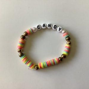 May include: A colorful beaded bracelet with the word "CANDY" spelled out in white beads. The bracelet is made with alternating gold beads and multi-colored beads.