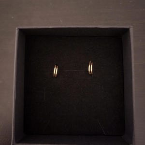 May include: A pair of small, gold-coloured hoop earrings presented in a black box. The earrings have a simple, circular design. The box is rectangular with a dark interior, creating a contrasting backdrop for the jewellery.