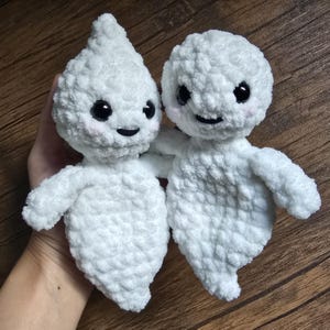 May include: Two white crocheted ghost plushies with black eyes and smiling mouths. The ghosts are holding hands and have a fluffy texture.