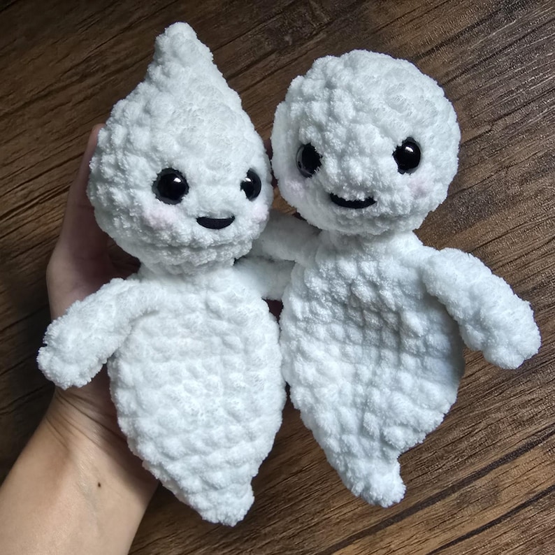 May include: Two white crocheted ghost plushies with black eyes and smiling mouths. The ghosts are holding hands and have a fluffy texture.