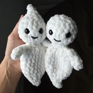 May include: Two white crocheted ghost plushies with black eyes and smiling mouths. The ghosts are holding hands and are being held by a hand.