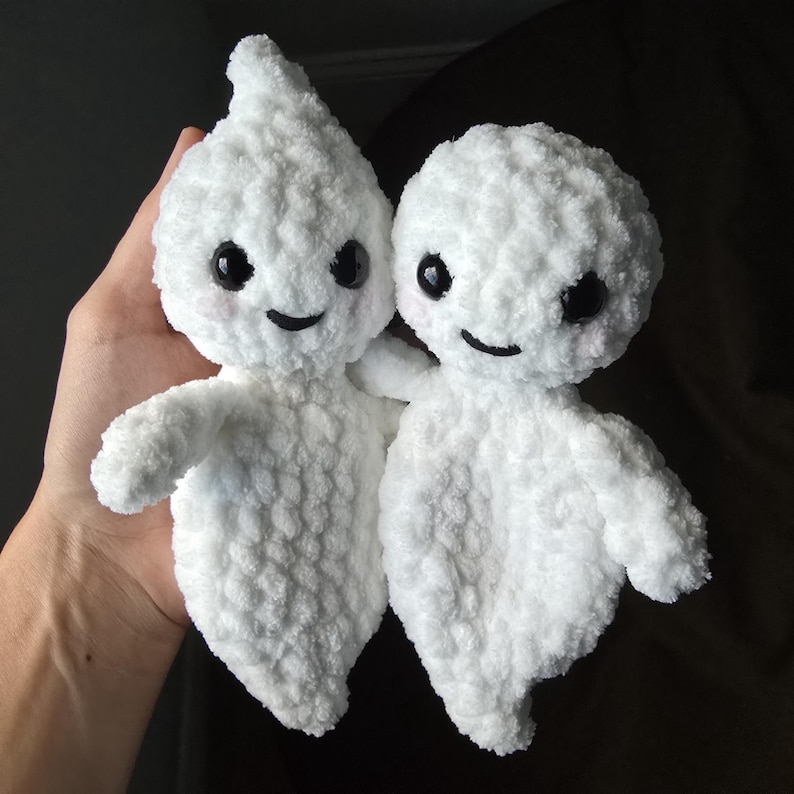 May include: Two white crocheted ghost plushies with black eyes and smiling mouths. The ghosts are holding hands and are being held by a hand.
