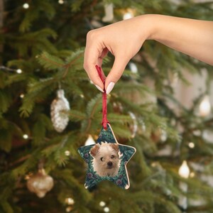 May include: Wooden star-shaped Christmas ornament featuring a dog's face surrounded by a green wreath design. The ornament is held by a red ribbon against a blurred Christmas tree background.