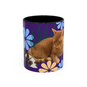 May include: A purple ceramic mug with a black rim features a stylized floral design with orange and blue flowers. A ginger cat is curled up on the mug, with a smaller white and brown cat peeking out from behind it.