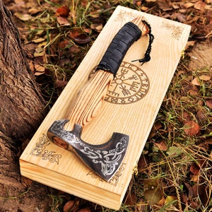May include: A hand-forged axe with a black leather handle and intricate engravings. The axe is displayed on a wooden board with a compass and runes.