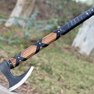 Puede incluir: Un hacha negra y marrón con un mango de madera envuelto en cuero negro. El mango está asegurado con remaches de latón. La cabeza del hacha tiene una hoja curva y una punta afilada.