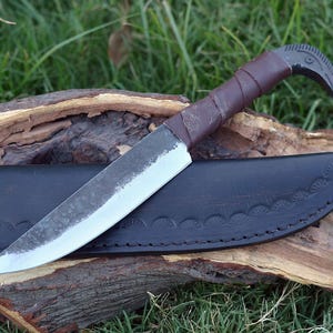 May include: A handmade knife with a curved blade and a brown leather handle. The knife is in a black leather sheath with decorative stitching.