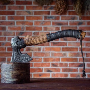 May include: A hand-forged axe with a wooden handle and a black leather wrap. The axe head is decorated with intricate carvings and a runic symbol. The axe is resting on a wooden log.