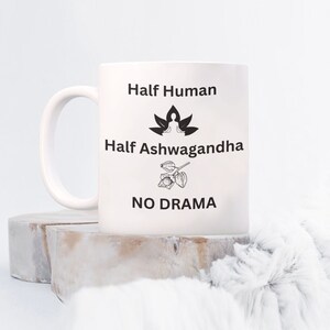 May include: White ceramic mug with black text that reads "Half Human, Half Ashwagandha, No Drama". The text is centered on the mug. There is a black and white illustration of a person meditating on a lotus flower above the text "Half Ashwagandha" and a black and white illustration of an ashwagandha plant below the text "No Drama".