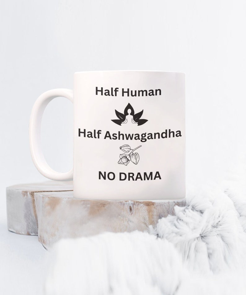 May include: White ceramic mug with black text that reads "Half Human, Half Ashwagandha, No Drama". The text is centered on the mug. There is a black and white illustration of a person meditating on a lotus flower above the text "Half Ashwagandha" and a black and white illustration of an ashwagandha plant below the text "No Drama".