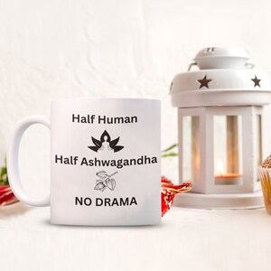 May include: White ceramic mug with black text that reads "Half Human Half Ashwagandha No Drama". The mug has a black illustration of a person meditating in a lotus position and a drawing of an Ashwagandha plant.