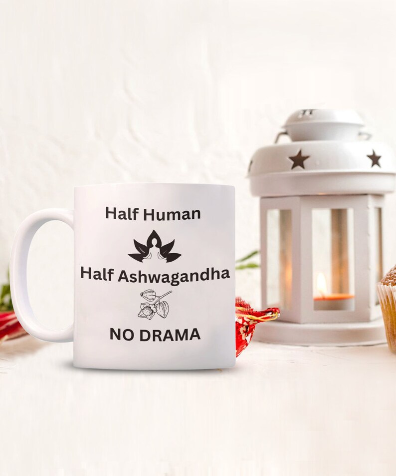 May include: White ceramic mug with black text that reads "Half Human Half Ashwagandha No Drama". The mug has a black illustration of a person meditating in a lotus position and a drawing of an Ashwagandha plant.