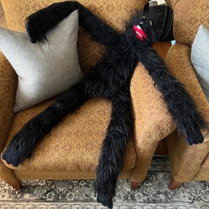 May include: A black furry puppet with a red mouth and black eyes is lying on a brown patterned armchair with a gray pillow. The puppet's arms are outstretched and its legs are crossed.