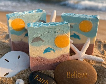 May include: Handmade soap bars with a beach theme, featuring a blue sky, yellow sun, and dolphin design. The bars are on a sandy beach with seashells, a starfish, and stones that say "Faith" and "Believe".