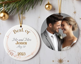 Könnte beinhalten: Zwei runde, weiße Keramik-Weihnachtsornamente. Eines zeigt ein Foto eines lächelnden Paares. Das andere hat den Text "Best of Mr and Mrs Jones Aug 30 2023" in goldener Schrift und ein goldenes Blumendesign. Beide hängen an goldenen Schnüren.