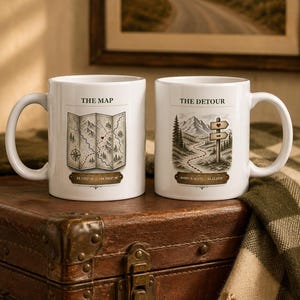 May include: Two white ceramic mugs featuring illustrations. The left mug displays a map with the text THE MAP and coordinates 39.7392 N 104.9903 W. The right mug shows a mountain path with the text THE DETOUR and AVERY & SCOTT 06.21.2019.