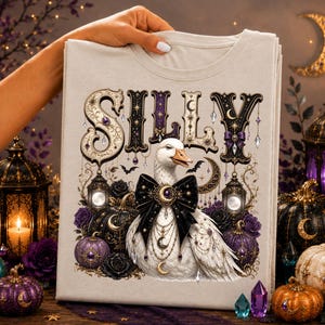 Pode incluir: Uma t-shirt bege claro com o gráfico de um ganso branco usando um grande laço preto e joias douradas com tema lunar. A palavra SILLY está impressa em letras decorativas escuras acima do ganso, rodeada por lanternas e abóboras.