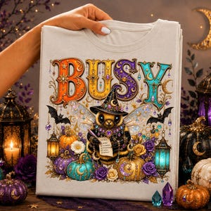 May include: A light gray t-shirt featuring a graphic of a bee wearing a witch hat and wings, surrounded by pumpkins, lanterns, and flowers. The word BUSY is printed in large, colorful, decorative letters above the bee, which holds a to-do list.