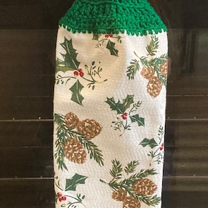 May include: A green crocheted kitchen towel holder with a brown button. The towel has a white background with a pattern of pine cones and holly.