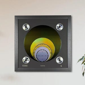 May include: A framed artwork featuring a circular design with concentric layers of yellow, green, and purple hues. The black frame has four silver-colored, circular accents. The artwork is mounted on a white wall.