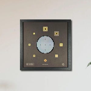 May include: A framed wall art piece with a dark brown frame. The artwork features a circular design in the center, surrounded by small gold squares and rectangles. The background is a dark brown color. The artwork is hanging on a white wall.