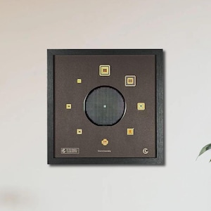 Puede incluir: Una obra de arte enmarcada con un fondo marrón oscuro y un marco negro. El arte presenta un diseño circular en el centro con un patrón de cuadrícula, rodeado de pequeños cuadrados y rectángulos dorados. La pieza está montada en una pared blanca.