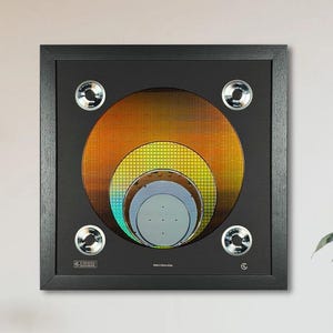 May include: Framed artwork featuring concentric circles in shades of orange, yellow, and blue, resembling a target or abstract design. The artwork is set against a black background and framed in a black wooden frame. Four silver-toned accents are placed around the artwork.