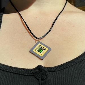 May include: A diamond-shaped pendant necklace with a black cord. The pendant has a gray border, gold inner frame, and a central green square. The necklace is worn around the neck.