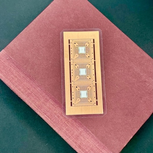 Op de afbeelding: Een rechthoekig, goudkleurig elektronisch component met drie vierkante microchips wordt weergegeven op een bordeauxrode ondergrond. De microchips hebben ingewikkelde interne ontwerpen en zijn ingekapseld in een transparante plastic hoes. De achtergrond is een gevouwen bordeauxrood papier.