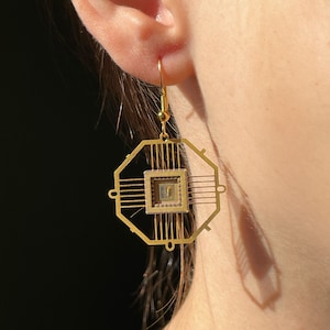 May include: Gold earrings with a geometric design featuring a small square in the center. The earrings are shaped like a circuit board.