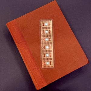Op de afbeelding: Een rechthoekige bladwijzer met een printplaatontwerp ligt op een gesloten, roodbruin boek. De bladwijzer heeft een transparante plastic hoes en heeft een gedetailleerd patroon van koperkleurige lijnen en kleine componenten. De kaft van het boek heeft een effen, getextureerde kleur.