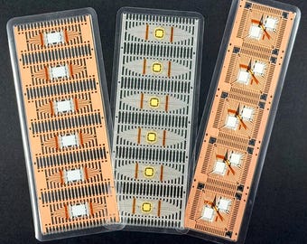 May include: Three rectangular circuit boards in clear plastic packaging. The boards have a copper-coloured base with black and gold circuitry and white square components. The boards are arranged on a black surface.