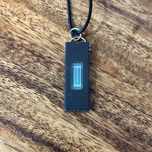 May include: A black rectangular pendant necklace with a silver-colored ring and a black cord. The pendant has a rectangular design with a light green and white illuminated center. The necklace is displayed on a wooden surface.