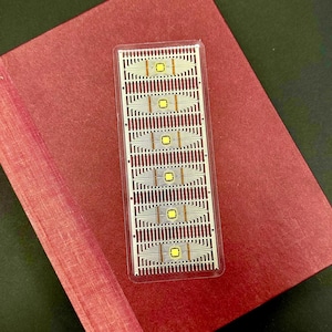 Op de afbeelding: Een rechthoekige elektronische component met een rasterpatroon van dunne, parallelle lijnen en kleine, vierkante gele elementen. De component is ingekapseld in een heldere plastic behuizing en rust op een rode ondergrond.