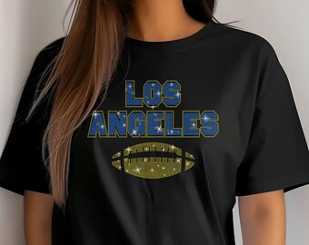 Puede incluir: Camiseta negra con el texto "LOS ANGELES" en azul y dorado, sobre un gráfico de balón de fútbol dorado. La camiseta es de cuello redondo y tiene mangas cortas.