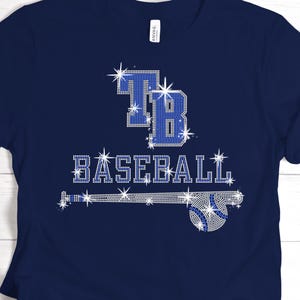 May include: Navy blue t-shirt with a rhinestone design. The design features the letters "TB" in blue rhinestones above the word "BASEBALL" in white rhinestones. A baseball bat and ball are also included in the design.