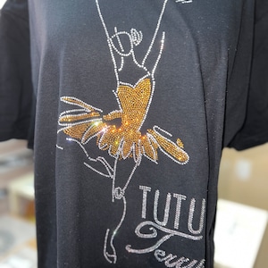 Peut inclure: T-shirt noir avec un motif en strass repr&eacute;sentant une ballerine en tutu. Le tutu est dor&eacute; et le contour de la ballerine est en strass. Le texte "TUTU Terry" est &eacute;galement en strass.