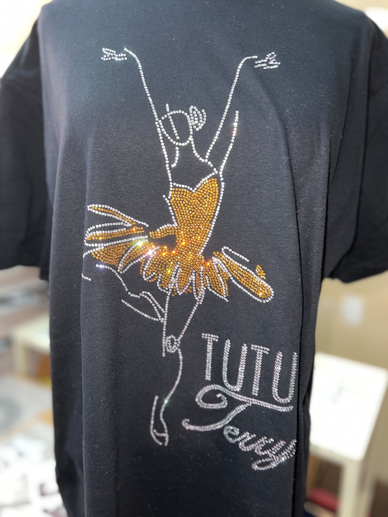 Peut inclure: T-shirt noir avec un motif en strass repr&eacute;sentant une ballerine en tutu. Le tutu est dor&eacute; et le contour de la ballerine est en strass. Le texte "TUTU Terry" est &eacute;galement en strass.