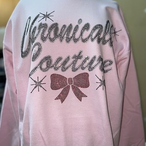 Op de afbeelding: Een lichtroze sweater met ronde hals en de tekst "Chronically Couture" in zilveren strass-steentjes op de borst. Onder de tekst staat een grote strik van rode strass-steentjes, geflankeerd door kleine sterretjes van zilveren strass-steentjes.