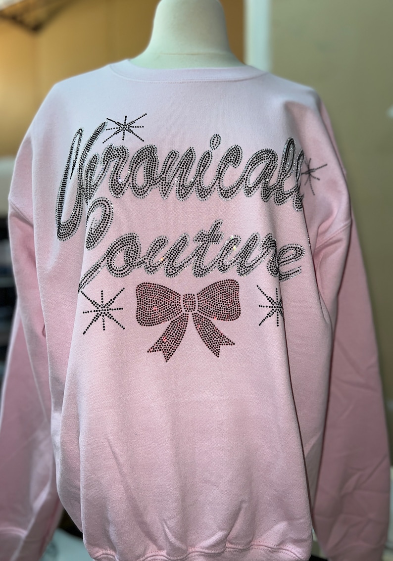 Op de afbeelding: Een lichtroze sweater met ronde hals en de tekst "Chronically Couture" in zilveren strass-steentjes op de borst. Onder de tekst staat een grote strik van rode strass-steentjes, geflankeerd door kleine sterretjes van zilveren strass-steentjes.