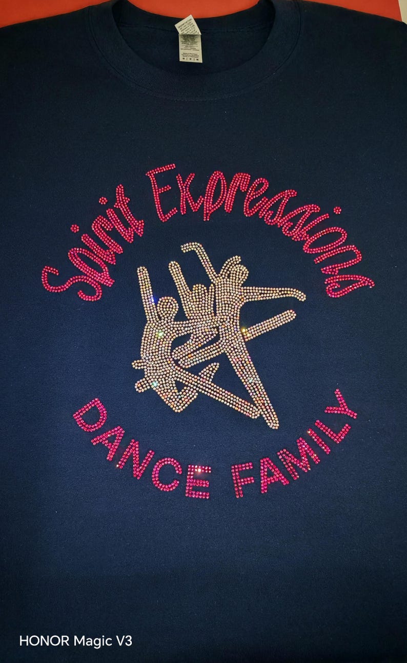 Op de afbeelding: Een marineblauw t-shirt met een strass-ontwerp van drie dansende figuren in het midden. De tekst "Spirit Expressions" is boven de figuren gebogen en "DANCE FAMILY" eronder, allemaal gemaakt van roze en doorzichtige strass-steentjes.
