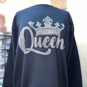 Op de afbeelding: Een zwart sweatshirt met lange mouwen en een groot grafisch ontwerp op de voorzijde. Het ontwerp bestaat uit het woord Queen in een cursief lettertype, bekroond met een gedetailleerde kroonillustratie, beide gemaakt met kleine, glinsterende zilveren strass-steentjes.