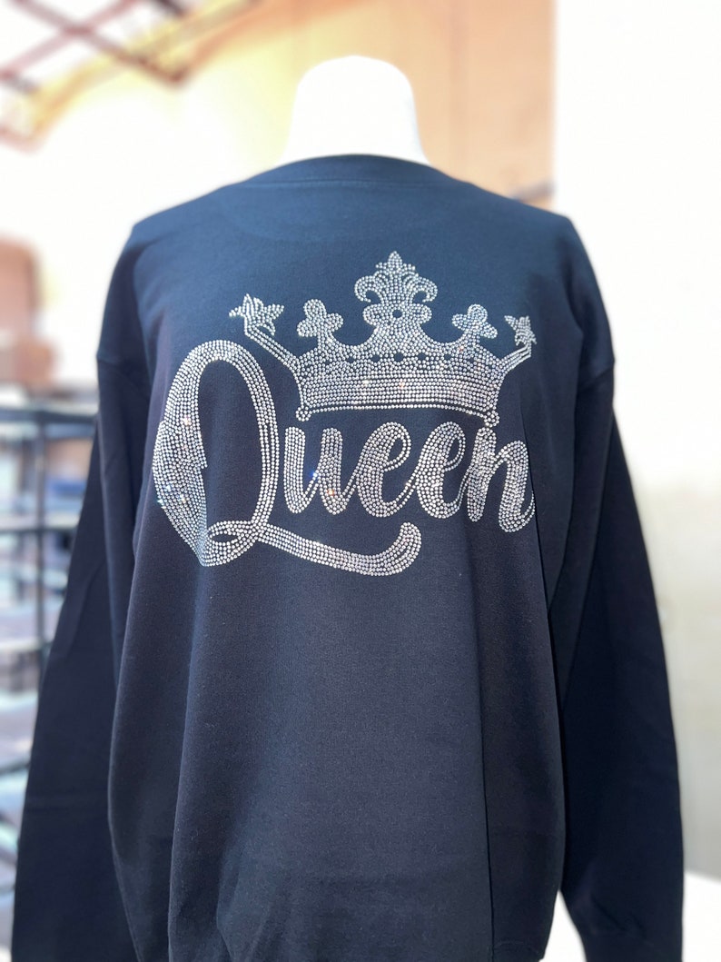 Op de afbeelding: Een zwart sweatshirt met lange mouwen en een groot grafisch ontwerp op de voorzijde. Het ontwerp bestaat uit het woord Queen in een cursief lettertype, bekroond met een gedetailleerde kroonillustratie, beide gemaakt met kleine, glinsterende zilveren strass-steentjes.
