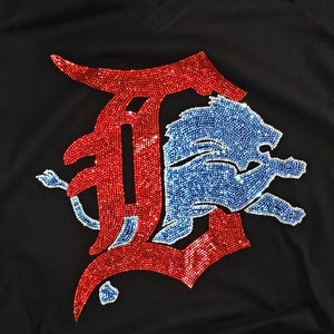 Op de afbeelding: Een zwart T-shirt met V-hals voorzien van een grote, decoratieve afbeelding gemaakt van rode en blauwe strass-steentjes. Het ontwerp bestaat uit een gestileerde rode letter D met daaroverheen een blauwe leeuwfiguur met glinstering.