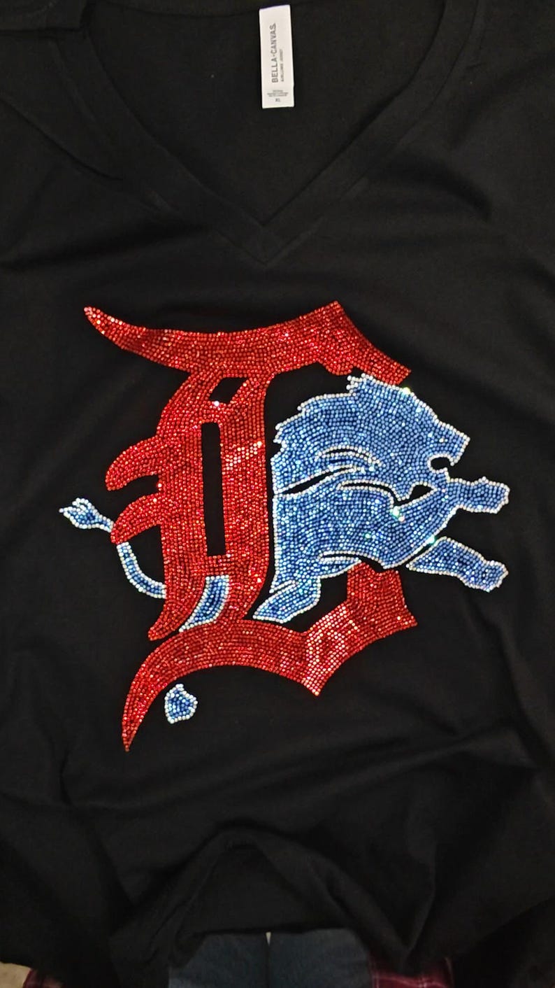 Op de afbeelding: Een zwart T-shirt met V-hals voorzien van een grote, decoratieve afbeelding gemaakt van rode en blauwe strass-steentjes. Het ontwerp bestaat uit een gestileerde rode letter D met daaroverheen een blauwe leeuwfiguur met glinstering.
